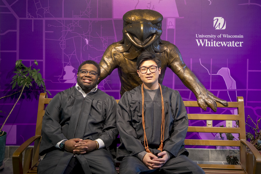 Two students wearing graduation gowns sit on a bench with a large metal Willie Warhawk behind them.