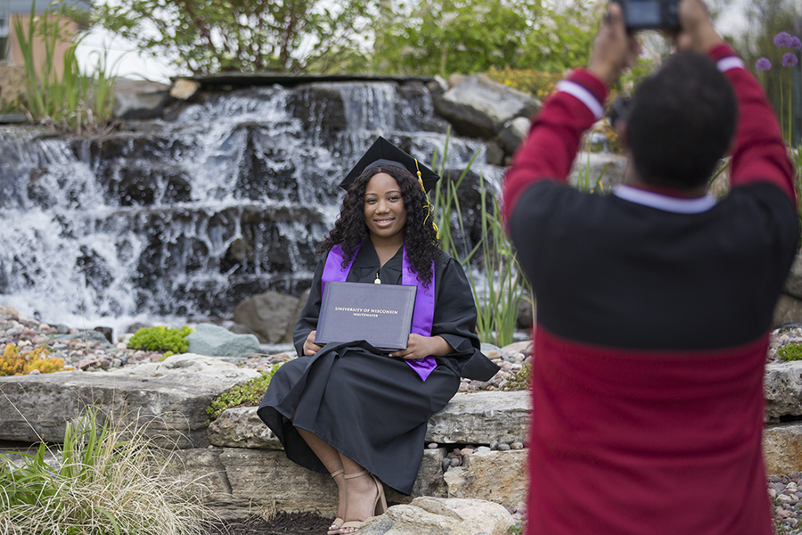 A student wearing graduation regalia and holding a diploma gets their picture taken in front a large water feature with flowing water.