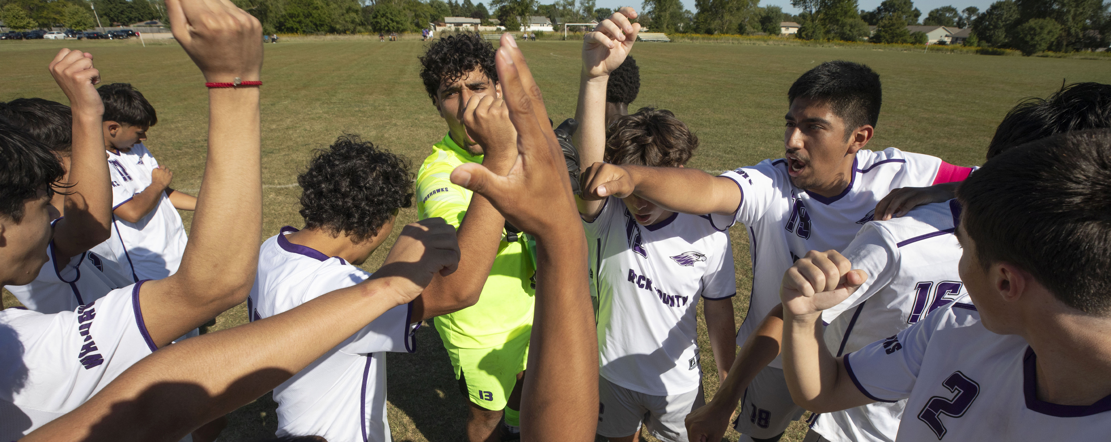 The men's soccer team gathers on the soccer field with their arms raised together in a huddle.