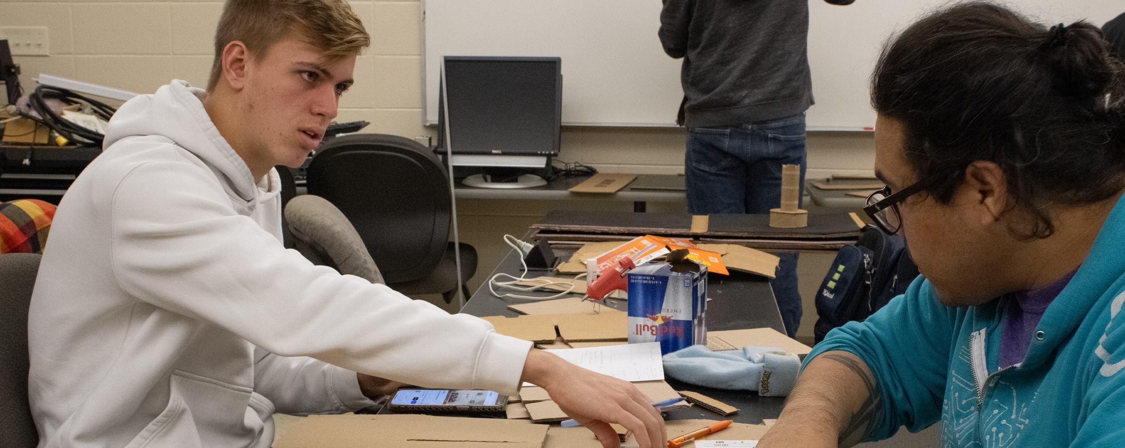 Two students work together at a table covered in cardboard pieces.