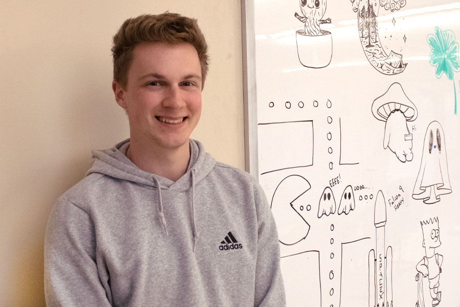 A student in a gray shirt stands next to drawings on a whiteboard and smiles at the camera.