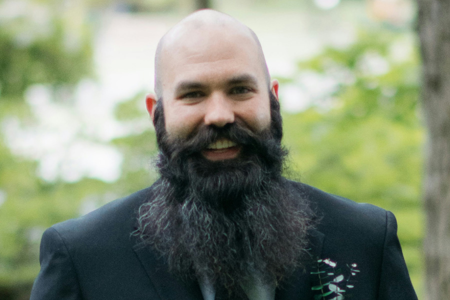 A person with a large beard wearing a black blazer smiles at the camera.
