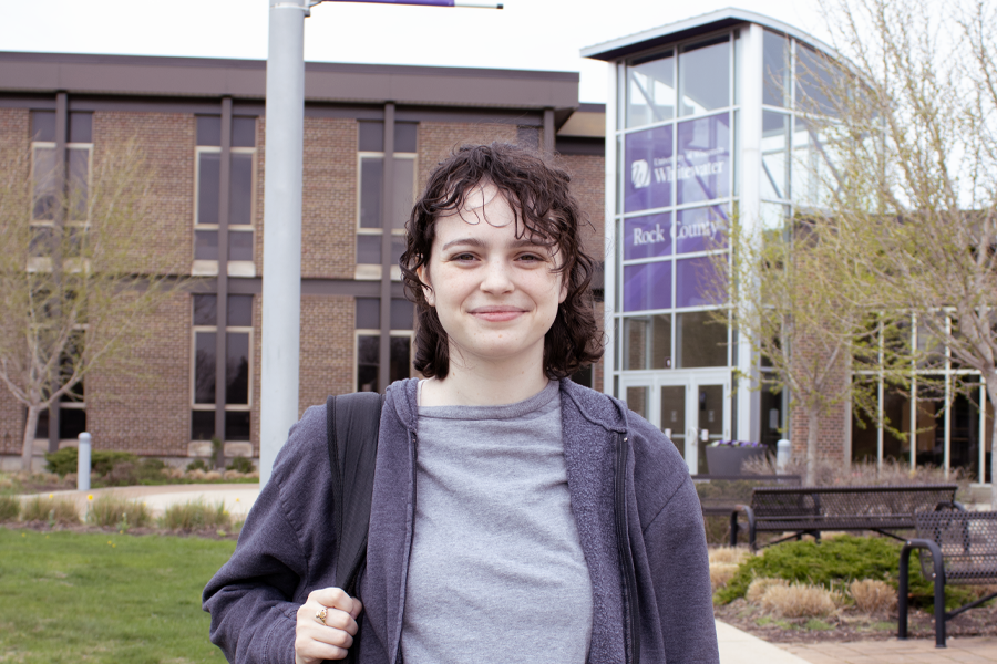 A person with a backpack over their shoulder stands outside of the main academic building on the Rock County campus smiles at the camera.