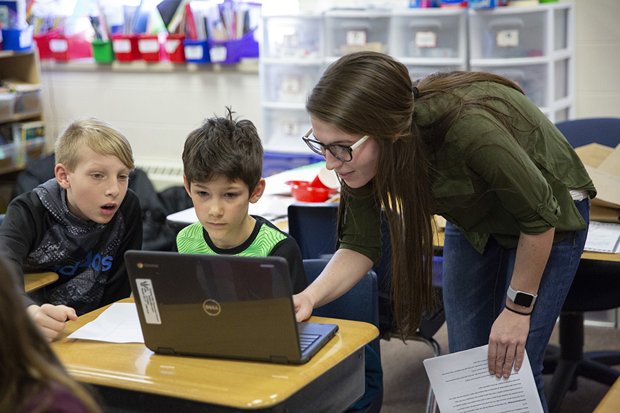 A student teacher wearing a green shirt leans over to assist two children on a laptop.