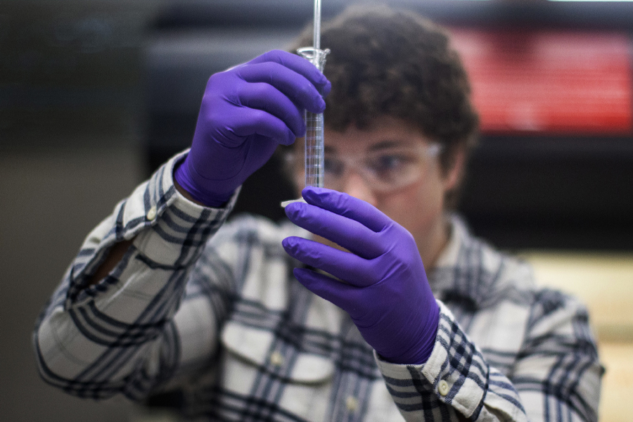 A student wearing purple rubber gloves and safety glasses inspects a test tube.