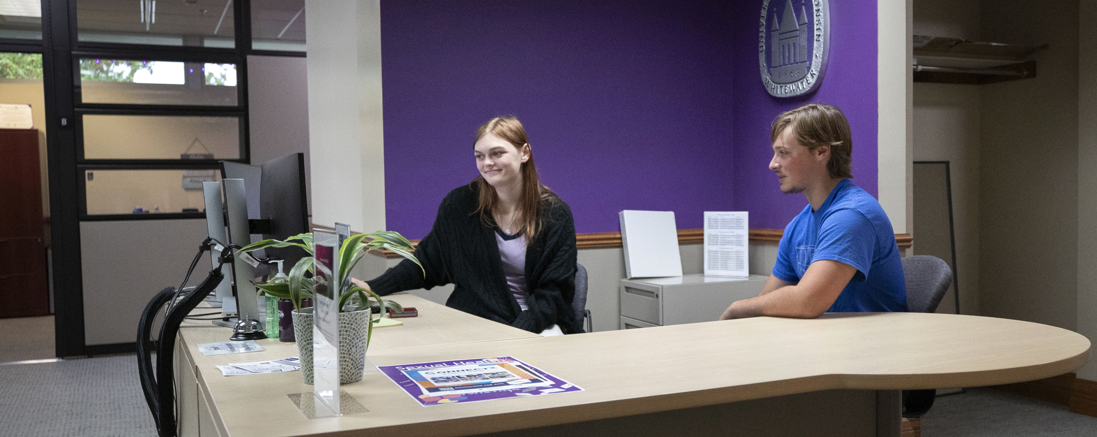 Two student work together at a desk in front of a purple wall.