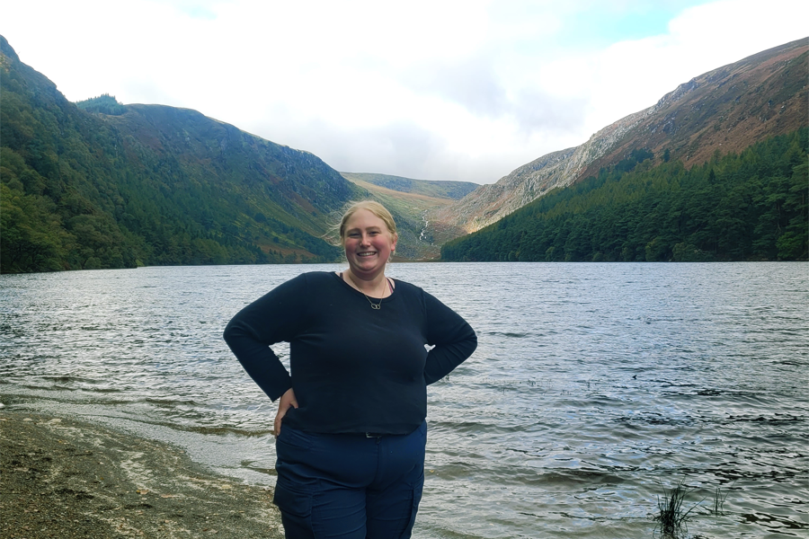 A student has their hands on their hips and a smile on their face as they stand in front of a lake surrounded by mountains. 