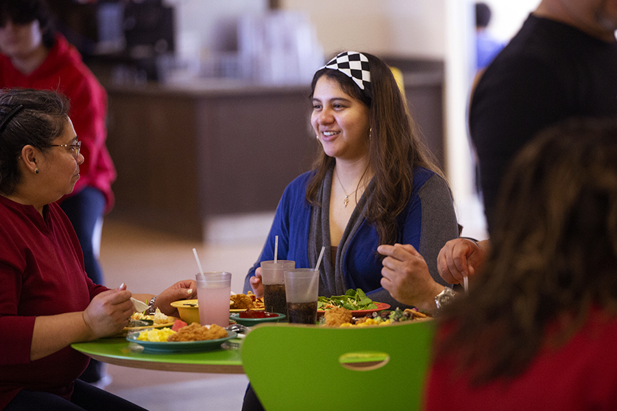 A group of people in a dining hall sit at a table with lime green chairs and several plates of food on the table.