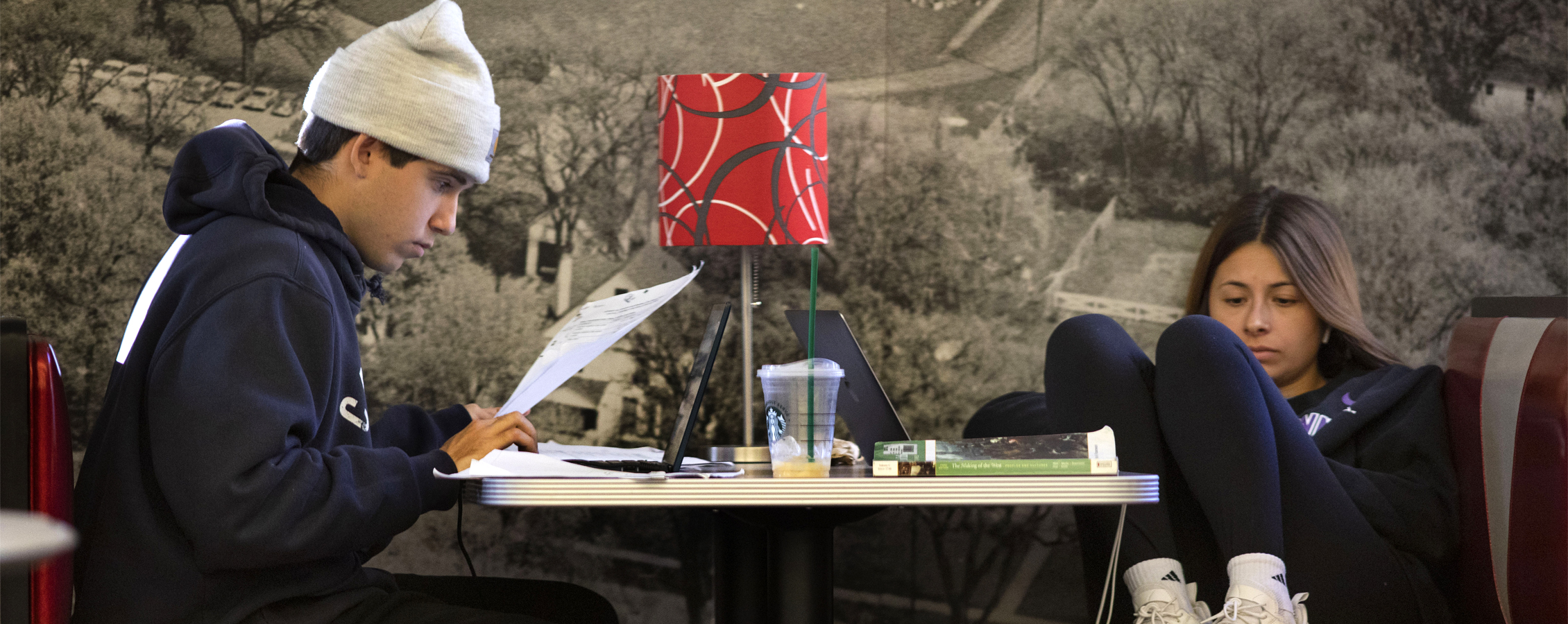 Two students study at a table with a red lamp on it.